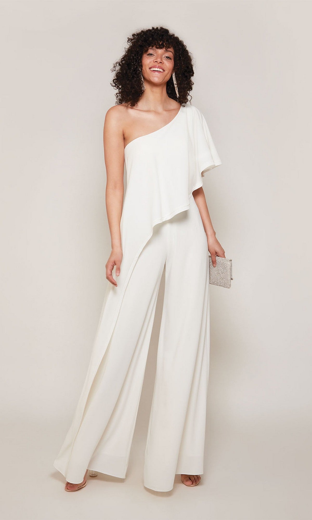 A woman poses against a plain background wearing the Alyce 70049 Long Formal Jumpsuit, a chic white one-shoulder outfit, and holds a small clutch bag.